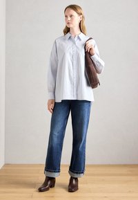 Frau steht drinnen und trägt ein hellblaues, oversized Hemd, hochgekrempelte blaue Jeans, braune Stiefeletten und hält eine braune Schultertasche.