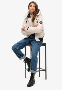 Padded, jasnoróżowa kurtka z kapturem, czarnymi akcentami i naszywką, noszona na kremowym swetrze i niebieskich podwiniętych dżinsach, w zestawie z czarnymi wiązanymi botkami.