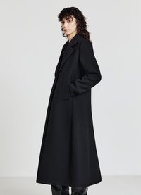 Cappotto lungo in lana nera con rever a punta, tasche laterali e silhouette dritta, che arriva a metà polpaccio. Texture liscia.