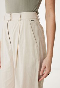 Beige hochgeschnittene Hose mit vorderen Falten, seitlichen Taschen und einem sichtbaren Markenlabel. Weicher Stoff mit einer dezenten Textur.