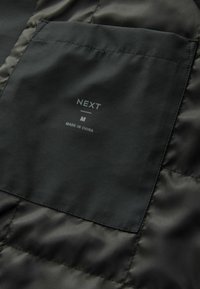 Tela de chaqueta acolchada negra con una textura suave; un bolsillo con parche etiquetado "NEXT", talla M, con "HECHO EN CHINA" debajo.