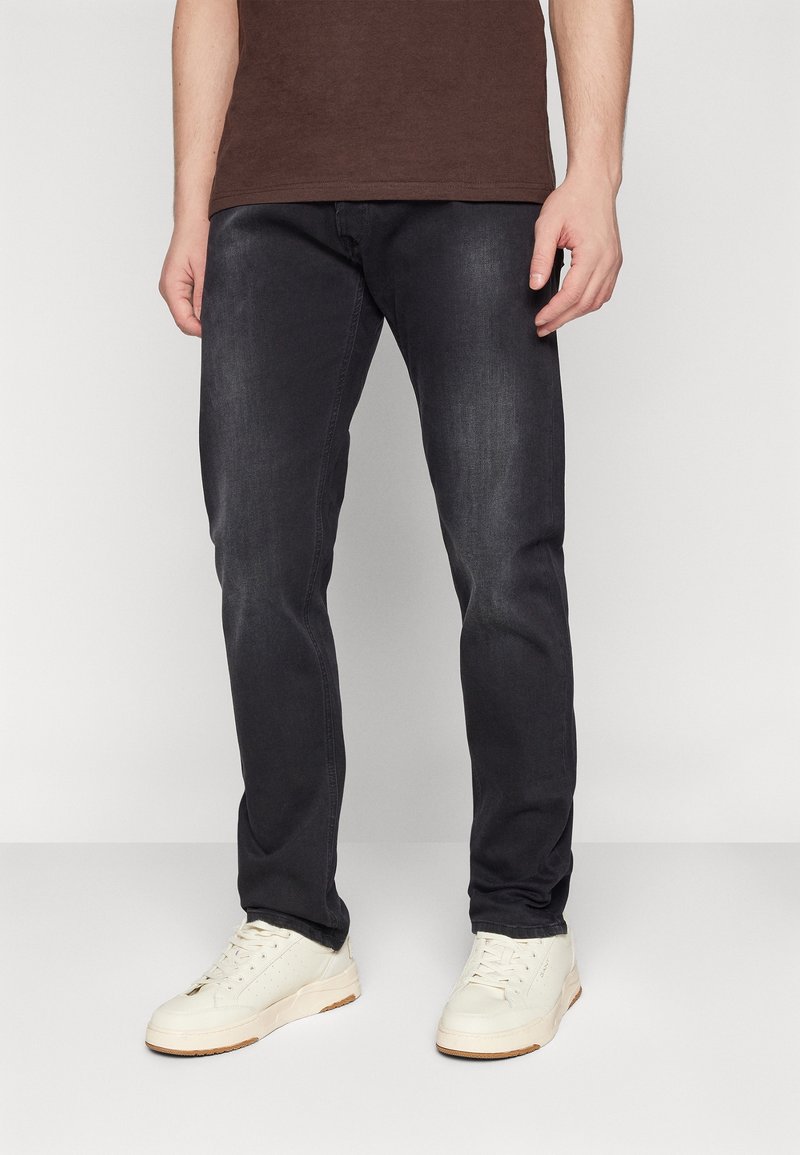 Homme portant un jean noir délavé, une chemise marron et des baskets blanches, debout devant un fond clair uni.