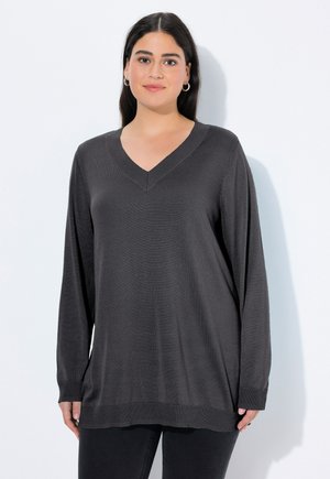 Femme aux longs cheveux noirs portant des boucles d'oreilles argentées en forme de créoles et un pull en V gris foncé ample sur fond blanc.