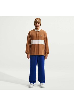 Mann trägt braun-weißes Langarm-Poloshirt, blaue Hose, weiße Sneaker und eine braune Mütze, steht vor einem einfarbigen hellen Hintergrund.