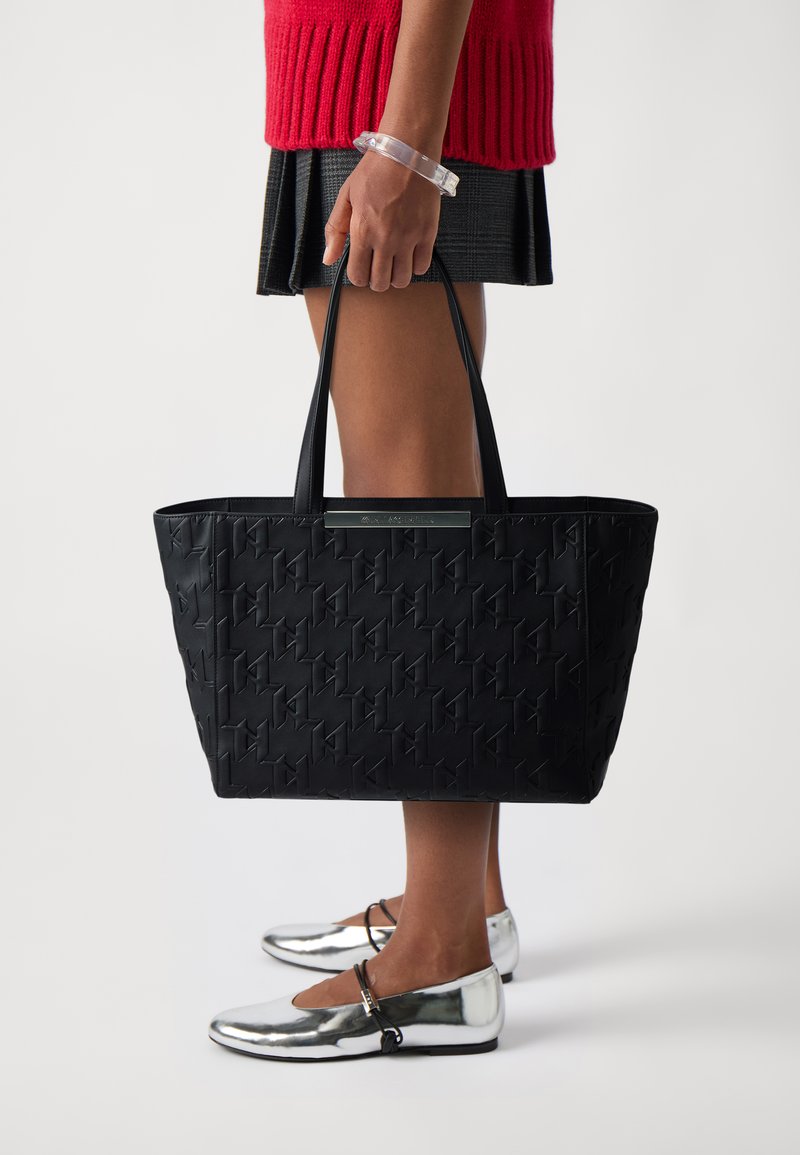 Borsa tote nera con texture, dotata di manici piatti, caratterizzata da un design a motivo e da un accento metallico sul fronte. Indossata con ballerine argentate.