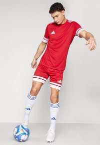 Röd atletisk tröja och shorts med vita ränder, kompletterat med blå och vita mönstrade strumpor och en flerfärgad fotboll.