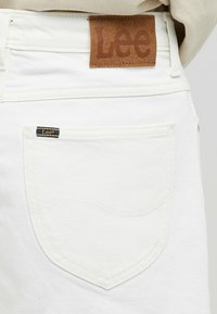 Bílé džíny z denimu s hnědou koženou značkou "Lee" na zadní části pasu a malou černou značkou na kapse. Jemné detaily šití.
