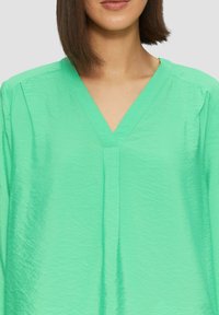 Muntgroene blouse met een V-hals, lichtgewicht stof, lichte textuur en plooidetail aan de voorkant. Korte mouwen, losse pasvorm.