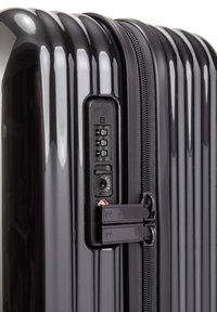Valigia rigida nera con scanalature verticali, chiusura con zip e lucchetto a combinazione. Presenta una finitura lucida e meccanismo di chiusura approvato TSA.