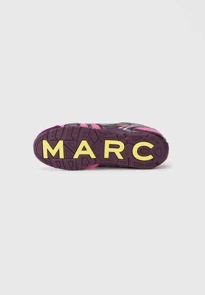 Semelle de baskets en violet et rose avec de grandes lettres jaunes écrivant "MARC" sur le dessous, contre un fond blanc uni.