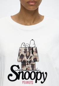 Άτομο που φοράει λευκό μπλουζάκι με τον Snoopy να лежει σε σκυλόσπιτο με καμουφλάζ σχέδιο και το κείμενο "Snoopy Peanuts" από κάτω.
