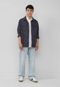 Marineblauwe button-up jas met twee borstzakken, gedragen over een witte polo, gecombineerd met lichtblauwe rechte jeans en sneakers.