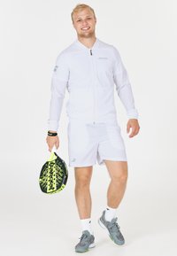 Ung mand i hvidt sportstøj, der holder en gul og sort Babolat padel racket, smilende og stående foran en helt hvid baggrund.