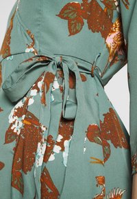 Vihreä kukkamekko pitkillä hihoilla, kapealla vyötäröllä, joka on sidottu kangasvyöllä. Ominaisuuksina ruskeat ja valkoiset kukkakuviot pehmeällä, teksturoidulla kankaalla.