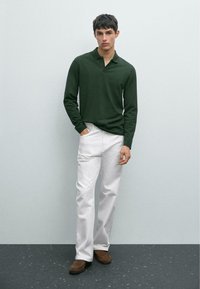 Donkergroen polo met lange mouwen, witte recht-toe-rechtaan broek en bruine suède schoenen. Model staat tegen een lichtgrijze achtergrond.