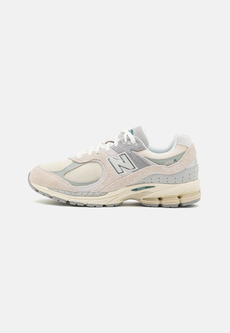 Baskets New Balance beige clair et gris avec panneaux en mesh et daim, lacets blancs, semelle rembourrée et logo "N" sur le côté.