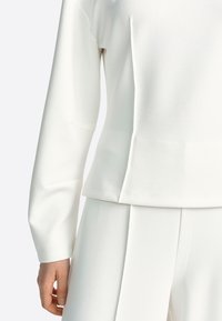 Valge pika varrukaga t-särk, millel on lahtine lõige, ümara kaelusega ja küljetasku detail, valmistatud siledast, venivast kangast.