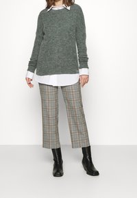 Grå stickad tröja över en vit skjorta, tillsammans med rutig byxor i brunt och svart, samt svarta ankelstövlar.