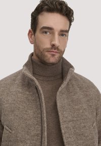 Braune Wollmischungjacke mit hohem Kragen und subtiler Textur, über einem passenden Rollkragenpullover in ähnlichem Farbton getragen.