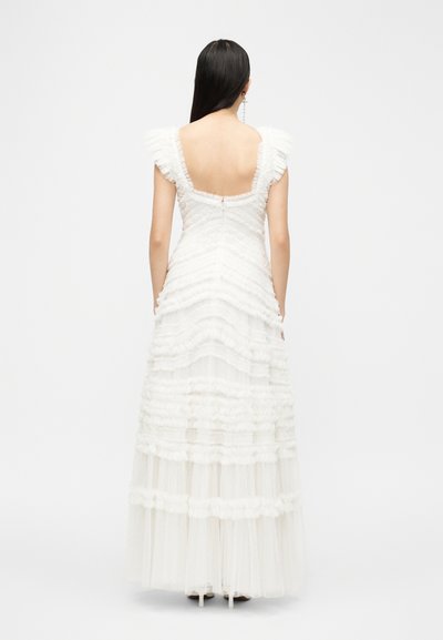 Γυναίκα με μακριά σκούρα μαλλιά που φοράει ένα λευκό φόρεμα από δαντέλα με στρώσεις, με σούρες στους κοντούς μανίκες και χαμηλή τετράγωνη πλάτη, στέκεται σε λευκό φόντο.