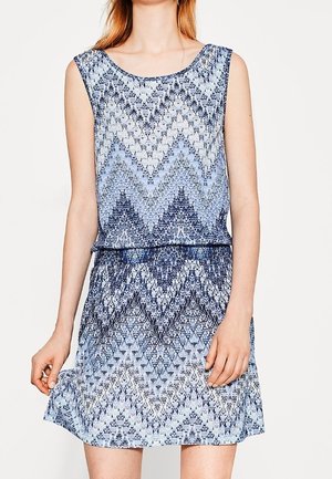 Mujer con un vestido sin mangas, de patrón azul y blanco con diseño de chevron, sujetando el dobladillo por un lado.