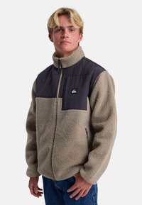Chaqueta de forro polar en beige con un panel en tela oscura en el pecho. Cuenta con un cierre con cremallera, cuello alto y parche con el logotipo en el bolsillo del pecho.