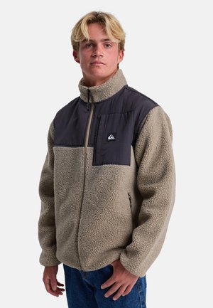 Fleece jacket in beige with a dark fabric chest panel. Features a zippered closure, high collar, and logo patch on chest pocket.