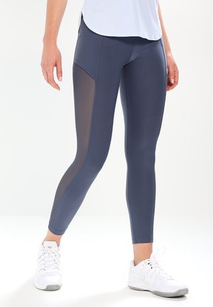 Frau trägt dunkelgraue Leggings mit schwarzen Mesh-Einsätzen an den Seiten und weiße Sportschuhe, steht vor einem einfarbigen Hintergrund.