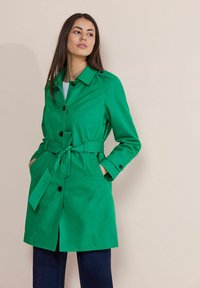 Grön trenchcoat med bälte i midjan, med spetsig krage och knappstängningar. Tillverkad av ett smidigt, lätt material.