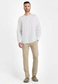 Man staat gekleed in een wit overhemd met lange mouwen, beige broek en bruine suède schoenen tegen een effen witte achtergrond.