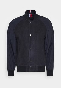 Chaqueta bomber azul marino con frente de ante, mangas de nailon, cierre de botones a presión, puños acanalados y cuello con detalle de rayas rojas y blancas.