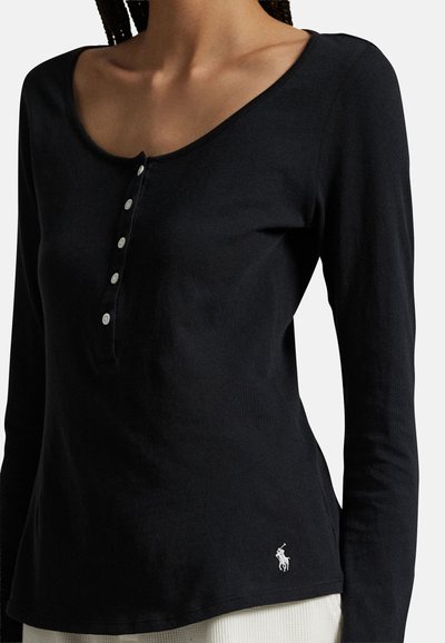 Femme portant un henley noir à manches longues avec cinq boutons blancs et un petit logo de joueur de polo blanc près de l'ourlet.