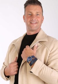 Beige wollen mix jack met zilveren rits; zwarte shirt eronder. Marineblauwe horloge met ronde wijzerplaat en gestructureerde band. Drie leren armbanden.