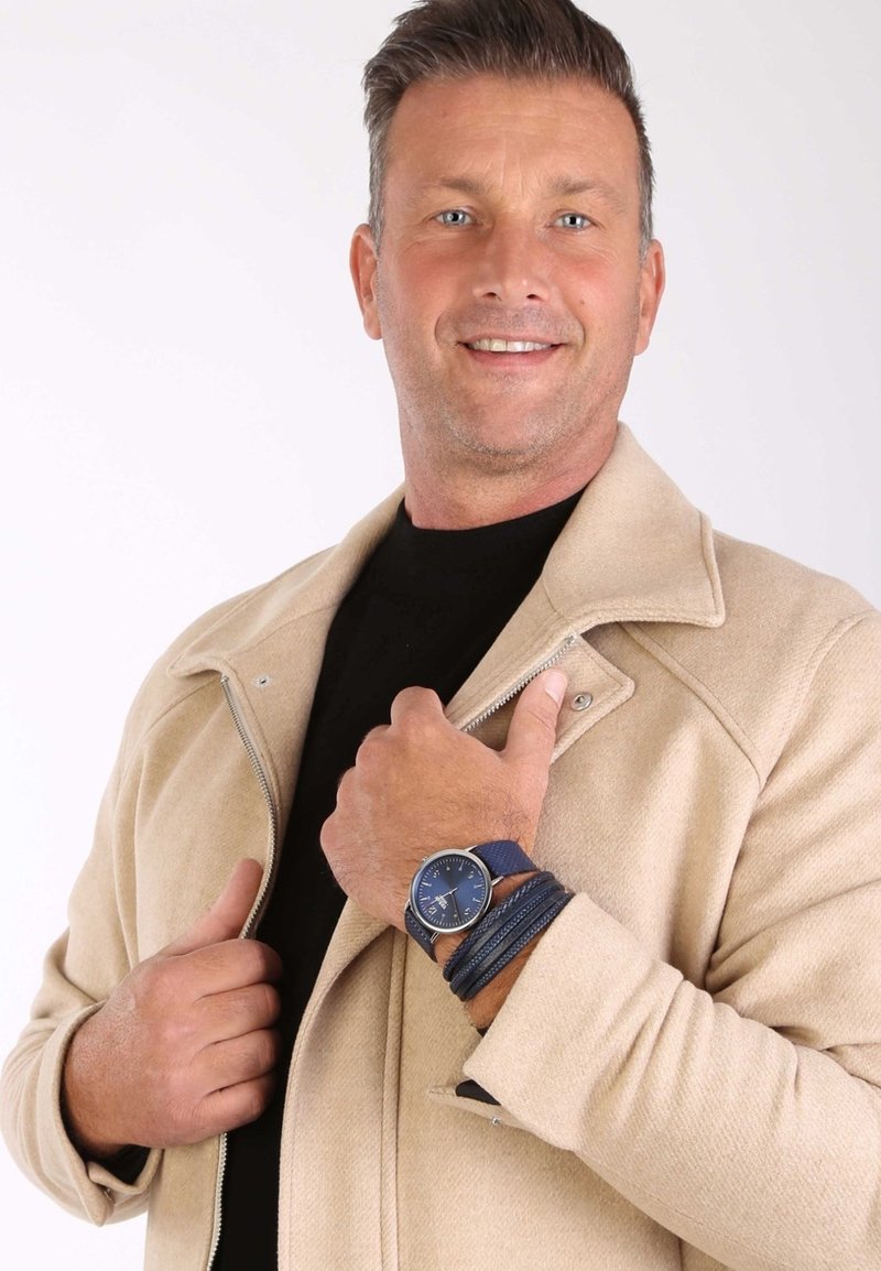 Beige wollen mix jack met zilveren rits; zwarte shirt eronder. Marineblauwe horloge met ronde wijzerplaat en gestructureerde band. Drie leren armbanden.