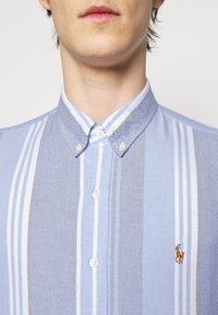 Ljusblå skjorta med knappar som har växlande smala och breda vita ränder, med en liten broderad logotyp på vänster bröst.