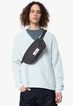 Zwarte heuptas met een ritssluiting en logo-patch, crossbody gedragen, gecombineerd met een lichtblauwe gestructureerde breisweater en donkere jeans.