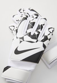 Vita och svarta målvaktshandskar med geometriska mönster, utrustade med en texturerad yta och en kardborreband för säker passform. Nike-logotypen är närvarande.