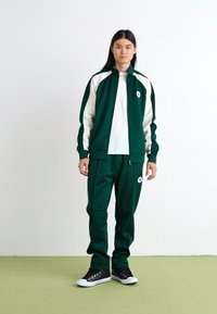 Πράσινο αθλητικό σακάκι με λευκές λεπτομέρειες και λογότυπο, συνδυασμένο με ταιριαστά πράσινα παντελόνια. Μαύρα αθλητικά παπούτσια προσθέτουν αντίθεση. Λείος, αθλητικός υφασμάτινος ιστός.
