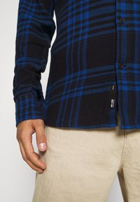 Camicia di flanella a quadri nera e blu con maniche lunghe, chiusura con bottoni e un'etichetta laterale, abbinata a pantaloni beige. Tessuto dalla texture morbida.