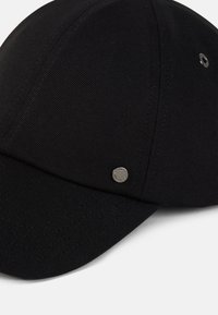 Black baseball cap with a textured fabric, round crown, and a silver metal accent on the side. Seam detailing is visible.