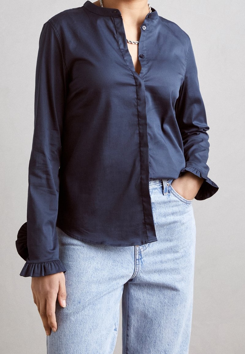 Blusa de manga larga color azul marino con cuello alto, botones al frente y puños con volantes, hecha de una tela suave. Combinada con jeans de mezclilla claros.