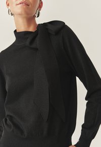 Femeie îmbrăcată cu un top negru cu mâneci lungi, având un fundă mare pe umăr și cercei argintii în formă de lacrimă, pe un fundal simplu.