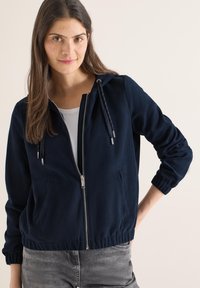 Sweat à capuche zippé de couleur marine avec cordon de serrage réglable, poches latérales et poignets élastiques. Fabriqué en tissu doux et texturé.