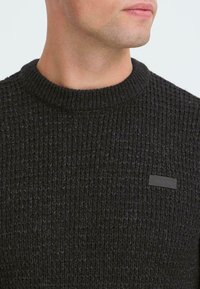 Sort strikket sweater med et tekstureret mønster, rund halsudskæring og et lille sort mærke i nederste venstre hjørne.