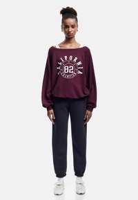 Sudadera de gran tamaño color burdeos con el gráfico "CALIFORNIA 82", pantalones de chándal negros y zapatillas blancas con una correa, de pie contra un fondo liso.