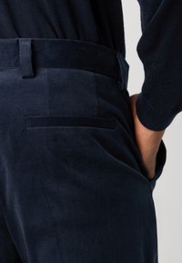 Un pantalon en velours côtelé bleu marine présente une texture douce, une coupe droite et une poche arrière avec une couture subtile. La taille est lisse et ajustée.