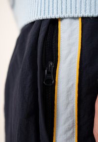 Juodas užtrauktukas artimame plane ant tamsiai mėlynų kelnių su baltais ir geltonais vertikaliais dryžiais, derinamas su šviesiai mėlynu megztu megztiniu.