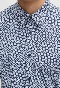 Námořnická modrá košile s krátkým rukávem, která má opakující se světle modrý květinový vzor, límec s knoflíky a tmavě modré knoflíky na přední straně.