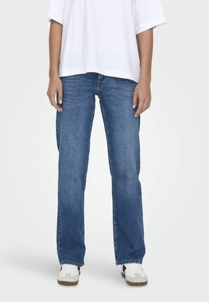 Jeans de mezclilla azul con un diseño de pierna recta, estilo de cinco bolsillos y una clásica cremallera de botones. Llevados con una camisa blanca de corte holgado y zapatillas deportivas.