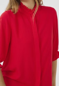 Blusa rossa con collo alto e dettagli arricciati, maniche a palloncino corte e chiusura con bottoni. Realizzata in tessuto leggero e morbido.